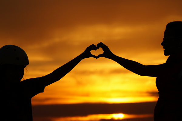 Silhouette of parent and child making heart shape with hands in front of sunset.