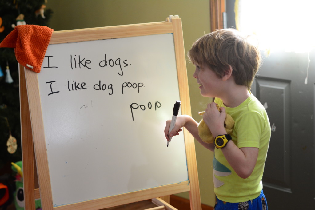 child copying words from a whiteboard. the whiteboard says "I like dogs. I like dog poop." the child wrote "poop".